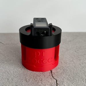 May include: A red and black device with the text "PT OCCC" on the side. The device has a small screen and the word "Alert" is visible. The device is cylindrical and appears to be 3D printed.
