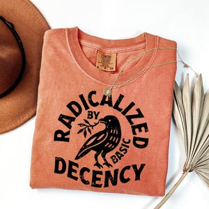 Op de afbeelding: Een koraalkleurig t-shirt met de tekst "RADICALIZED BY BASIC DECENCY" en een zwarte ravenafbeelding. Een gouden ketting en een bruine hoed zijn ook zichtbaar. Het shirt is gemaakt van Comfort Colors-materiaal.
