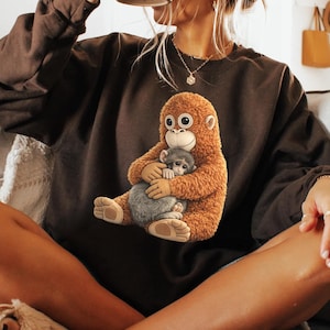 Puede incluir: Sudadera marrón con un dibujo de un orangután y un mono gris. El orangután es marrón con la cara blanca y ojos grandes. El mono gris está en los brazos del orangután.