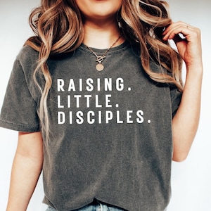 Könnte beinhalten: Dunkelgraues T-Shirt mit dem Text "RAISING. LITTLE. DISCIPLES." in weißen Blockbuchstaben. Das Shirt hat kurze Ärmel und einen Rundhalsausschnitt. Das Model trägt eine goldene Halskette.
