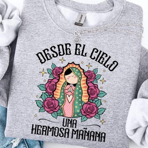 May include: A light gray sweatshirt featuring a graphic of a religious figure surrounded by pink roses and the text "DESDE EL CIELO UNA HERMOSA MAÑANA". A silver necklace with a pendant is also visible.