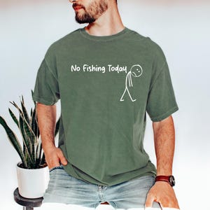 May include: Olive green t-shirt with the text "No Fishing Today" and a stick figure graphic. The shirt is a short-sleeved, casual style. The text and graphic are in white.