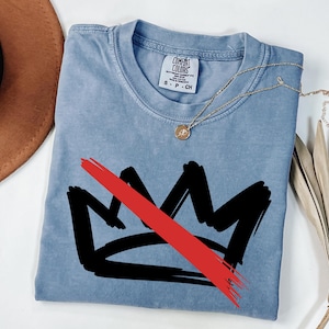 May include: A light blue Comfort Colors t-shirt featuring a black crown design with a red slash through it. A gold necklace with a pendant is draped across the shirt. The shirt is folded on a white surface.