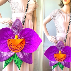 Puede incluir: Un bolso de mano de fieltro de color morado con forma de flor de orquídea con una correa de cebra en blanco y negro. El bolso de mano tiene un centro naranja brillante con un patrón de puntos.
