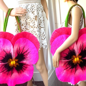 Puede incluir: Un bolso grande de fieltro rosa brillante con forma de flor de pensamiento. La flor tiene un centro negro con detalles amarillos y tallos verdes.