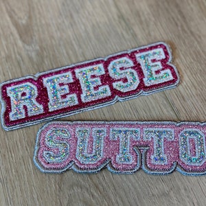 May include: Two pink and silver glitter iron-on patches with the names "REESE" and "SUTTON" in a varsity style font.