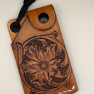 Puede incluir: Un tarjetero de cuero marrón con un diseño floral en relieve. El tarjetero tiene un cierre de botón a presión negro y un lazo de cordón negro. El diseño floral está grabado en la parte delantera. Costuras blancas visibles.