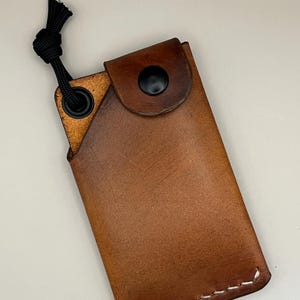 May include: A brown leather sheath with a black button closure and a black cord loop. The sheath is rectangular with white stitching along the bottom edge. The leather has a natural, textured appearance.