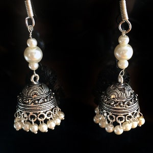 May include: A pair of silver earrings with white pearl accents. The earrings feature a bell-shaped design with intricate details and a dangling pearl drop.