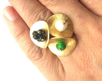Può includere: Un anello d'oro con tre pezzi di forme diverse. Il pezzo centrale ha una perla bianca, il pezzo a sinistra ha perline nere e il pezzo a destra ha una pietra verde.