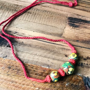 May include: A red cord necklace with three round ceramic beads. The beads are decorated with a floral pattern in shades of yellow, green, and brown.