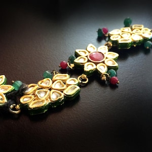 Op de afbeelding: Een goudkleurig haarstuk met groene en rode edelstenen. Het haarstuk bestaat uit verschillende bloemvormige stukken die met een ketting aan elkaar zijn verbonden.