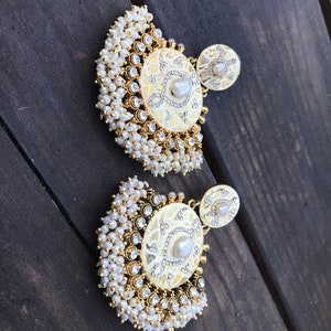 May include: A pair of gold earrings with white pearl accents. The earrings feature a round, white, decorative design with a pearl in the center. The earrings are surrounded by a fringe of white pearls.