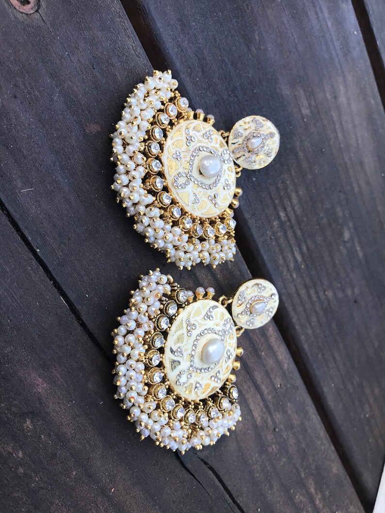 May include: A pair of gold earrings with white pearl accents. The earrings feature a round, white, decorative design with a pearl in the center. The earrings are surrounded by a fringe of white pearls.