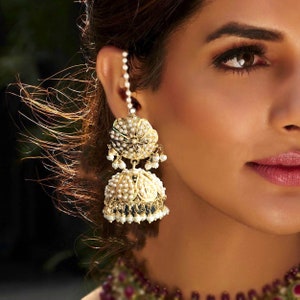 May include: A pair of gold and white jhumka earrings with pearl accents. The earrings are dangling and have a round shape. The earrings are worn by a person with dark hair.