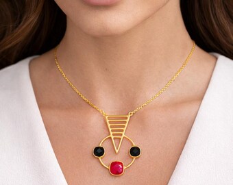 May include: A gold-tone necklace featuring a geometric pendant with a triangular top section and a circular base. The pendant is accented with two small round black stones and one larger square red stone set in the center of the circular frame.
