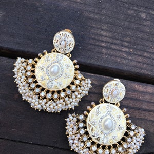 May include: A pair of gold earrings with white enamel and pearl accents. The earrings feature a round design with intricate details and a fringe of pearls.