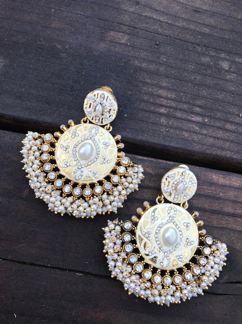 May include: A pair of gold earrings with white enamel and pearl accents. The earrings feature a round design with intricate details and a fringe of pearls.