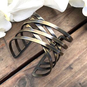 May include: A wide brass cuff bracelet with a woven design. The bracelet is made of multiple thin strips of metal that are interwoven to create a geometric pattern.