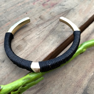 May include: A black and gold bracelet with a braided cord and metal accents. The bracelet is on a wooden surface.