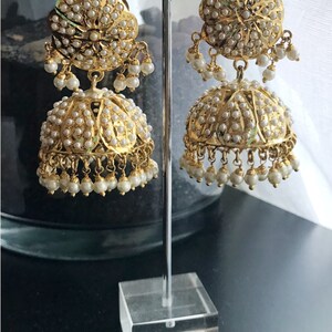 May include: A pair of gold earrings with pearl accents. The earrings are in the shape of a bell with a round, floral-shaped top. The earrings are displayed on a clear stand.