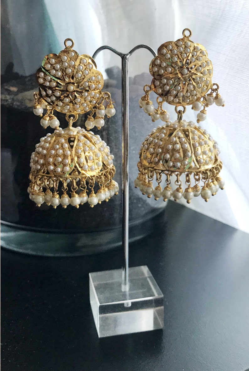 May include: A pair of gold earrings with pearl accents. The earrings are in the shape of a bell with a round, floral-shaped top. The earrings are displayed on a clear stand.