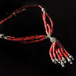 May include: A red coral necklace with a tassel pendant. The necklace is made of small, rectangular beads and silver spacers. The tassel is made of the same beads and has a silver bead at the end.