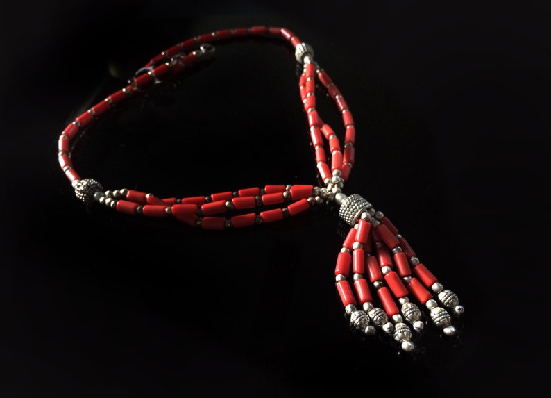 May include: A red coral necklace with a tassel pendant. The necklace is made of small, rectangular beads and silver spacers. The tassel is made of the same beads and has a silver bead at the end.