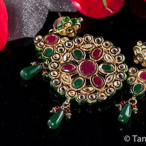 May include: A pair of gold earrings featuring green and red gemstones. The earrings are crafted from metal and have a traditional Indian design.