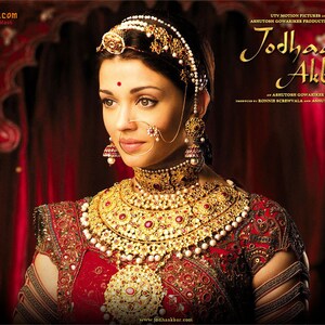 May include: A woman wearing a red and gold outfit with intricate embroidery and jewelry. She is wearing a large gold necklace, earrings, a nose ring, and a headpiece. The background is a red curtain. The text "Jodhaa Akbar" is visible in the upper right corner of the image.