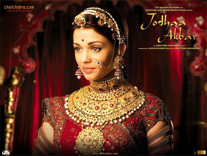 May include: A woman wearing a red and gold outfit with intricate embroidery and jewelry. She is wearing a large gold necklace, earrings, a nose ring, and a headpiece. The background is a red curtain. The text "Jodhaa Akbar" is visible in the upper right corner of the image.
