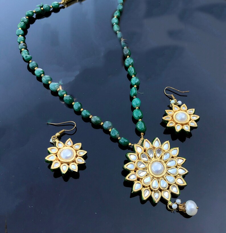 May include: A green gemstone beaded necklace with a large gold pendant featuring a white pearl surrounded by smaller white stones. Matching earrings with a white pearl center and smaller white stones.
