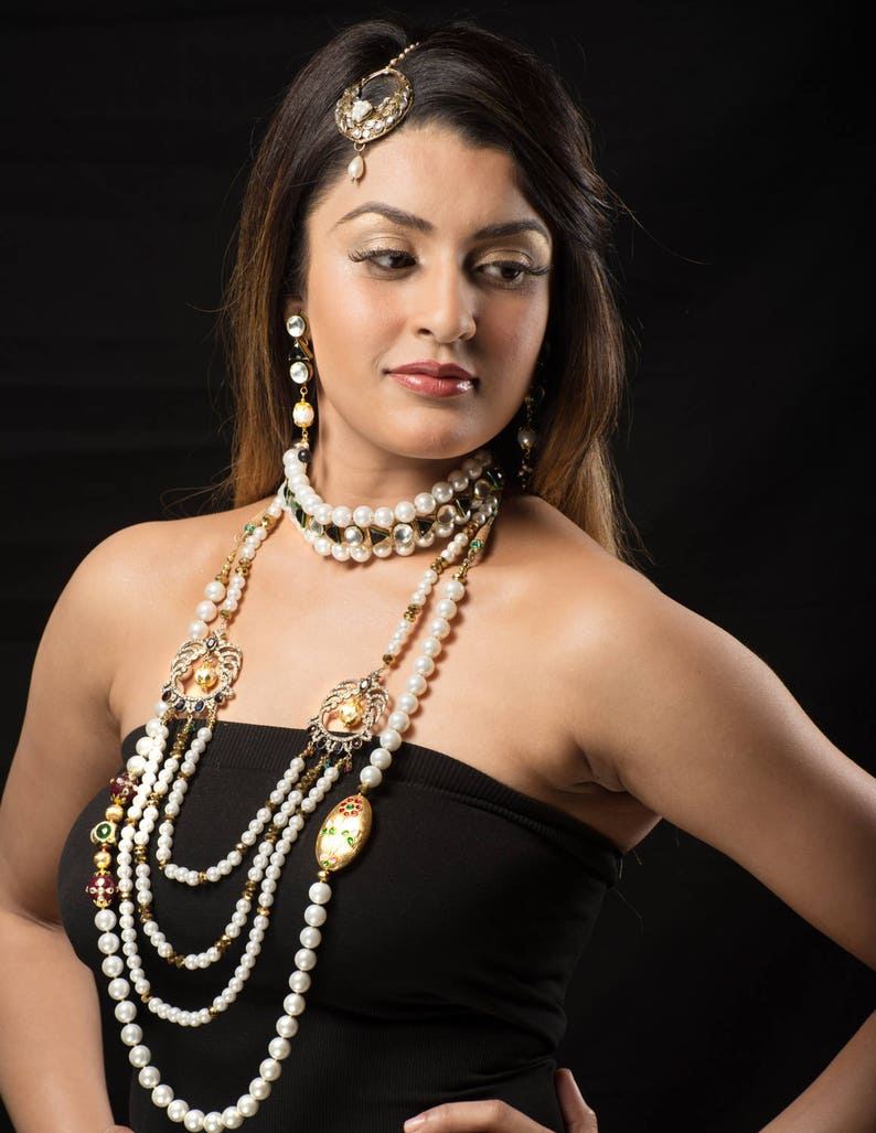 May include: A woman wearing a black strapless top and a layered pearl necklace with gold accents. The necklace features multiple strands of pearls and decorative pendants.
