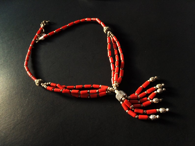 May include: A red coral necklace with silver beads and a tassel. The necklace is made of three strands of coral beads and has a silver clasp.