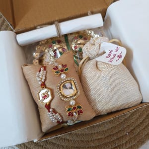 Puede incluir: Una caja de regalo de cartón que contiene dos pulseras decorativas sobre un cojín de tela beige, una pequeña bolsa de cordón beige con una etiqueta que dice Rakhi Ritual Kit y adornos metálicos dorados adicionales. Las pulseras cuentan con perlas blancas, hilo rojo y engastes de piedra centrales con detalles en rojo y verde.