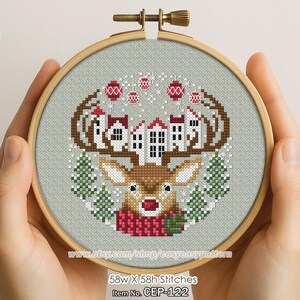 Op de afbeelding: Een kruissteekontwerp in een houten hoepel toont een rendier met geweien versierd met huizen. Het rendier draagt een rode sjaal, omringd door kerstboom- en ornamentmotieven. Het ontwerp staat op een lichtgrijze stof.