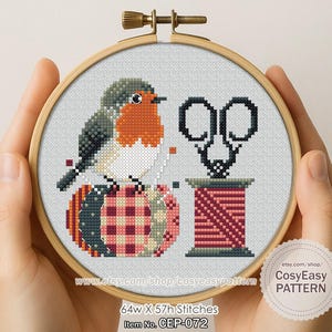 Könnte beinhalten: Kreuzstich-Stickerei in einem Holzreifen mit einem pixeligen Rotkehlchen, das auf einer Garnrolle sitzt. Schwarze Schere und eine rote Garnrolle befinden sich neben dem Vogel. Der Text "CosyEasy PATTERN" ist sichtbar.