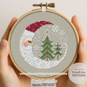 May include: A cross-stitch design in a wooden hoop features a crescent moon with Santa Claus's face and a red hat. Two green Christmas trees and snowflakes are also stitched. The design is held by hands.