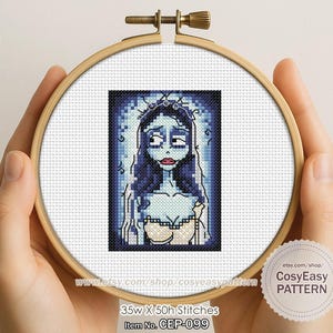 以下が含まれることがあります： 木製の刺繍枠に収められた、青い肌に長い濃い青色の髪、白いドレスと王冠を身に着けた女性のクロスステッチ刺繍。 「CosyEasy PATTERN」の文字が見えます。 パターンは幅35ステッチ、高さ50ステッチです。