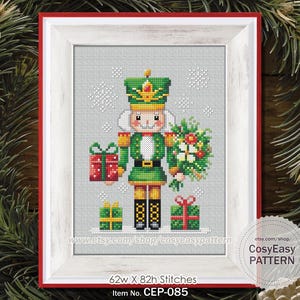 May include: Cross-stitch pattern of a Christmas nutcracker soldier in a white framed picture. The nutcracker is green, gold, and white, holding gifts and a bouquet. The frame is red and white. The text reads "62w X 82h Stitches" and "Item No. CEP-085".