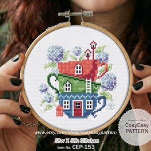 Op de afbeelding: Een kruissteekpatroon in een houten borduurring met een stapel van drie theekopjes ontworpen als huisjes. De rode, groene en blauwe kopjes zijn versierd met ramen, deuren en ladders, omringd door paarse en groene bloemclusters.