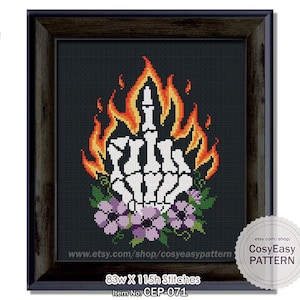May include: Cross-stitch pattern framed in a dark wood. The design features a white skeletal hand giving the middle finger, surrounded by flames in shades of orange and yellow, with purple flowers at the base. The text reads "CosyEasy PATTERN".