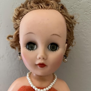 May include: A doll with light skin, green eyes, and short, curly light brown hair. The doll wears a white faux pearl necklace and matching faux pearl stud earrings. It is dressed in a strapless orange velvet top with a soft, textured finish.