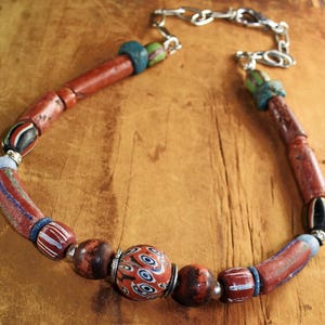 Puede incluir: Un collar artesanal con cuentas rojas, marrones, azules y blancas. Incluye cuentas cilíndricas y redondas, unidas por una cadena plateada. Las cuentas presentan patrones y texturas únicos, creando una pieza original.