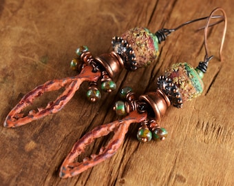 May include: A pair of dangle earrings featuring textured, rust-colored metal leaf-shaped charms, copper-toned bell caps, and multi-colored green and brown speckled beads. The earrings are finished with dark copper-toned wire hooks for hanging.