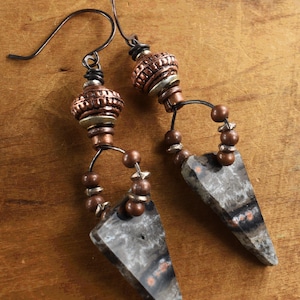 May include: A pair of dangle earrings with a triangular shaped stone pendant, copper beads, and a wire loop. The stone is a dark gray with brown and white streaks.
