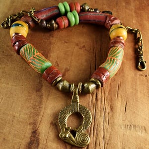 May include: A handmade necklace with a brass pendant and a beaded strand. The necklace features colourful beads in red, green, yellow, and brown. The pendant has an open-circle design. The necklace is displayed on a wooden surface.