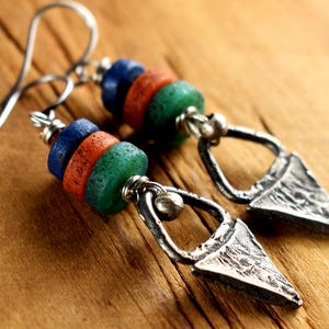 May include: A pair of handcrafted earrings featuring a combination of blue, orange, and green beads. The earrings have a silver-tone metal design with a textured, triangular pendant. The ear wires are also silver-toned.