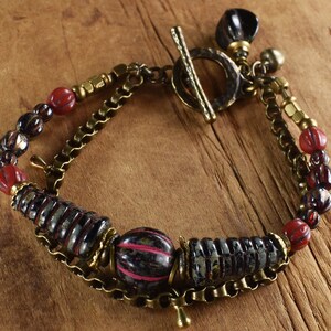 Puede incluir: Un brazalete de múltiples hilos con una cadena de latón y un cierre de palanca. El brazalete presenta cuentas de vidrio rojas y negras, incluyendo algunas con un patrón de rayas.