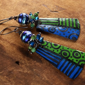 Puede incluir: Un par de pendientes con patrones geométricos azules y verdes en colgantes de metal. Los colgantes están unidos a un alambre de plata con cuentas de vidrio azul iridiscente y cuentas de cristal verde y azul.